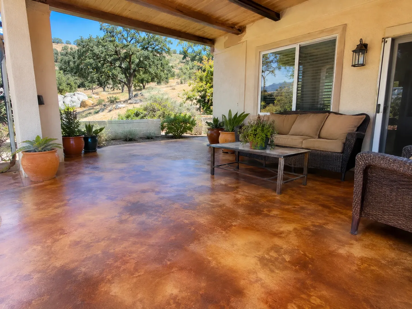 A covered patio with an acid-stained concrete floor in mottled amber, brown and caramel tones, sealed with a soft sheen, with wicker furniture, potted plants and a view of oak-dotted hills.