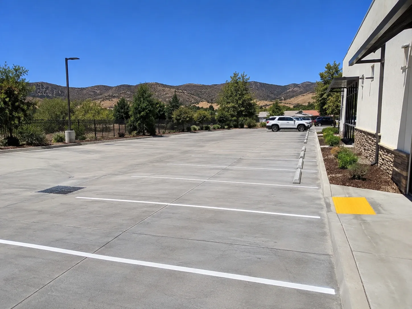 A new concrete parking lot with fresh white stall striping, saw-cut expansion joints, a drain inlet and wheel stops beside a commercial building, with dry hills behind.