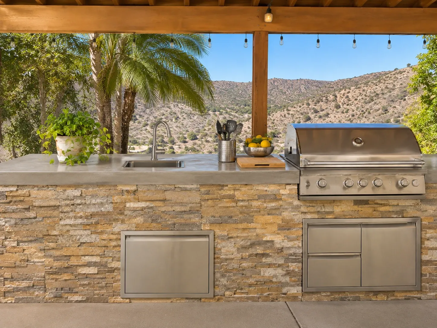 An outdoor kitchen with a long sealed grey concrete countertop with a square edge, a built-in stainless grill and sink on a stone-veneer base, under a covered patio overlooking dry hills.