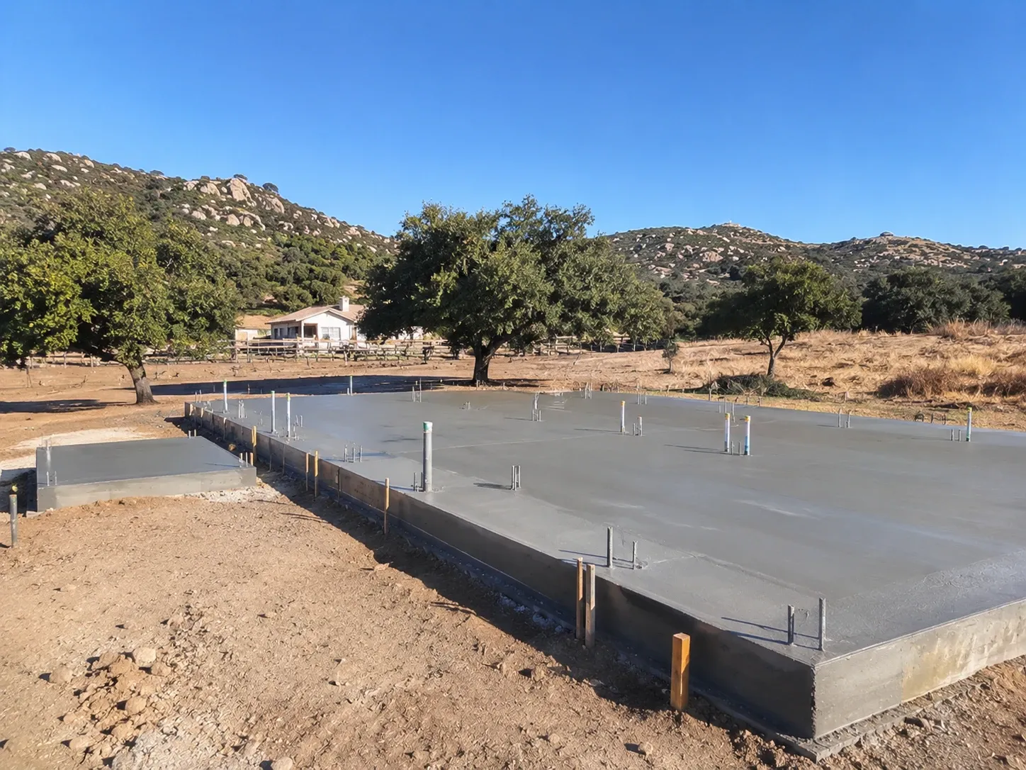 A freshly poured concrete foundation slab with thickened edges, conduit sleeves and anchor bolts on a rural lot, with a separate equipment pad, oak trees and boulder-strewn hills behind.