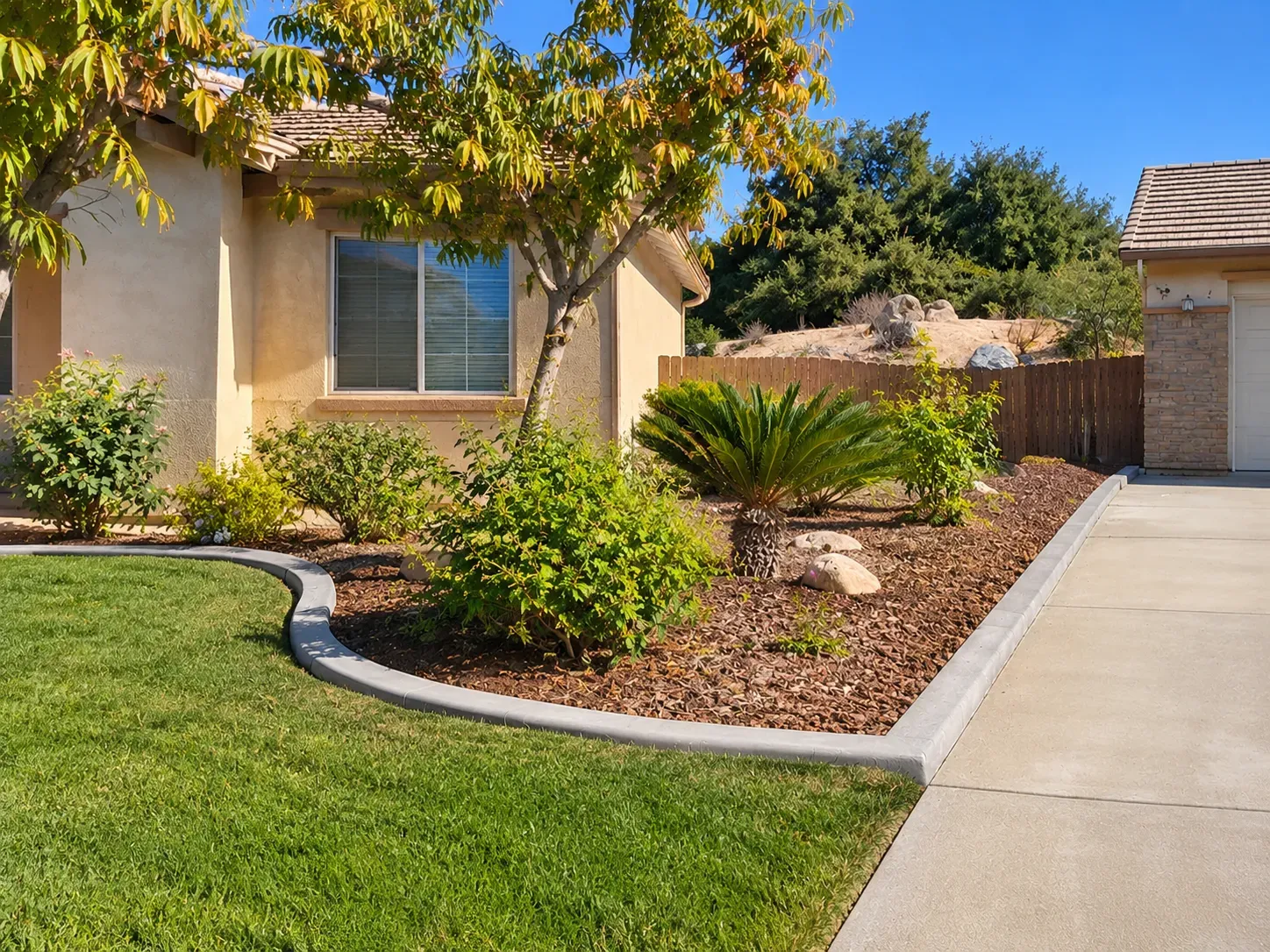 A smooth concrete landscape curb curving between a green lawn and a mulched planting bed, then running straight along the driveway of a stucco home.