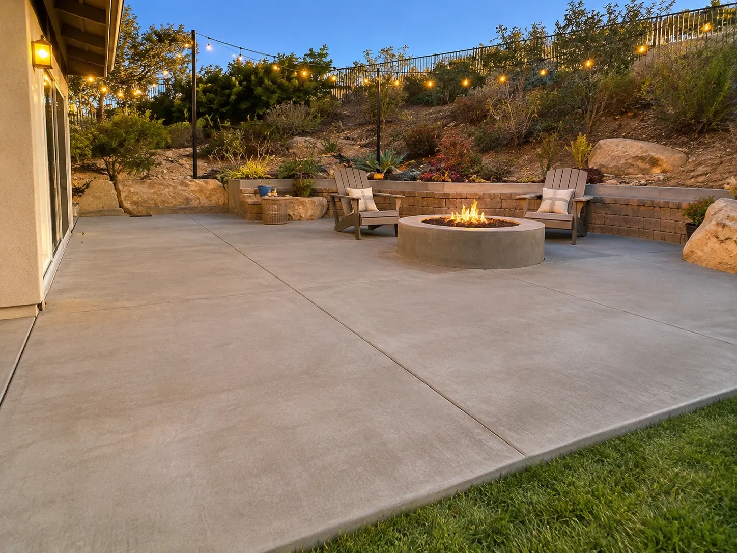 A large new concrete patio with saw-cut joints and a round concrete fire pit, outdoor chairs and string lights at dusk, below hillside planting behind a low block wall.