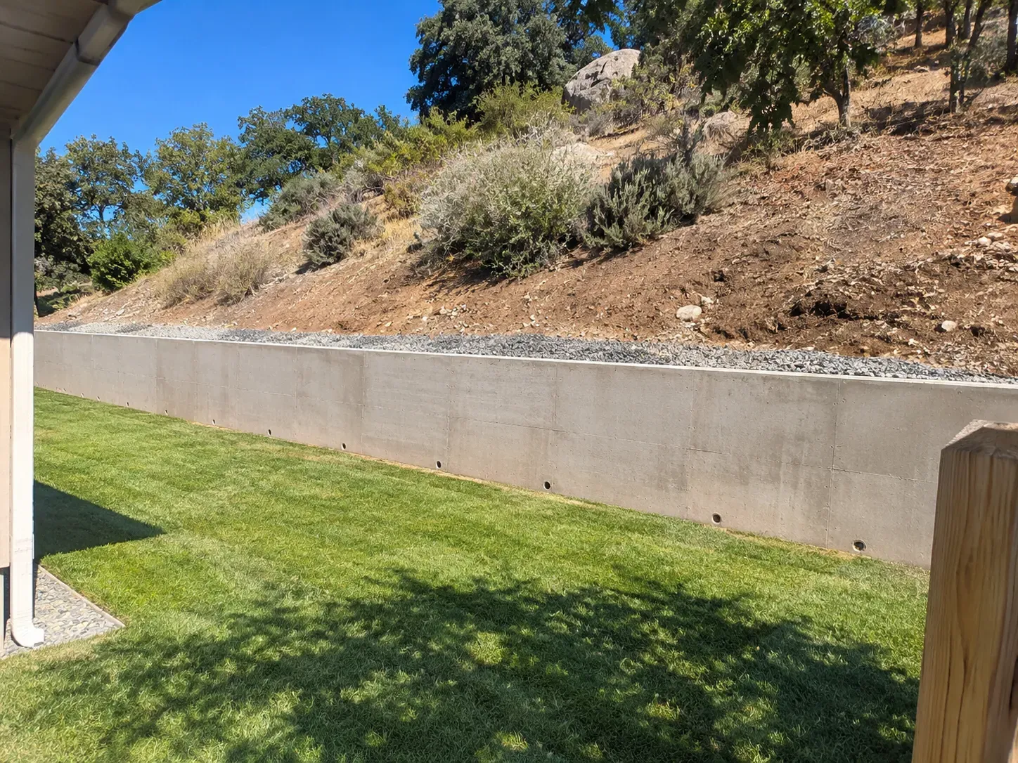 A poured concrete retaining wall holding back a hillside, with weep holes along its base, drainage gravel along the top and a new lawn in front.