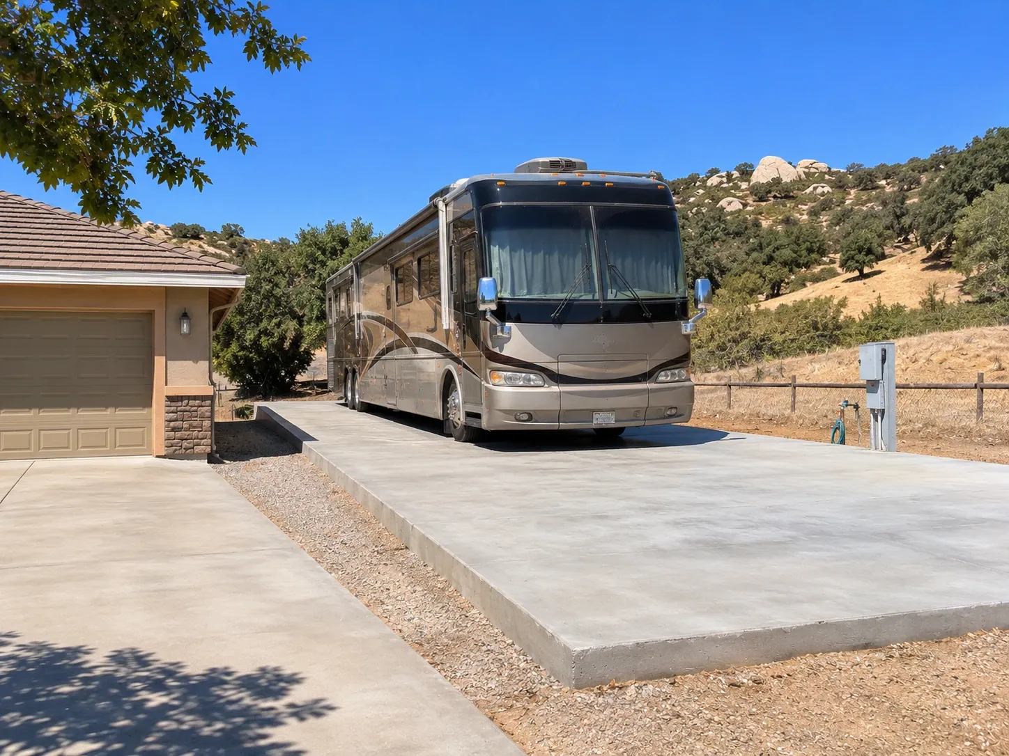 A motorhome parked on a thick new concrete RV pad beside a ranch home, with a power pedestal and water hookup at the pad edge and boulder-strewn hills behind.