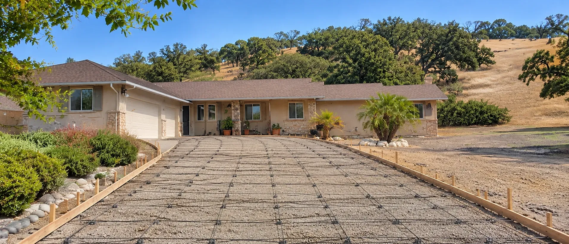 A residential driveway formed and ready for a concrete pour, with a gravel base, a rebar grid on chairs and wooden forms, in front of a ranch-style home below oak-covered golden hills.