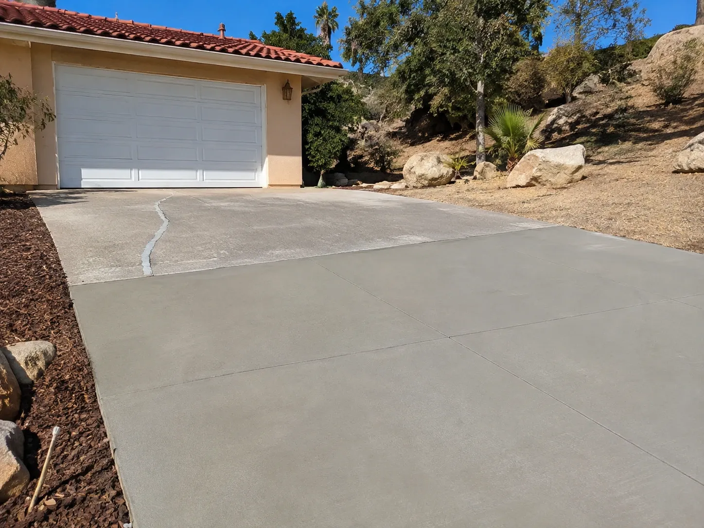 A driveway partway through repair: the lower section freshly resurfaced in an even grey finish with crisp joints, the upper section old concrete with a filled crack, below a stucco garage with a tile roof.