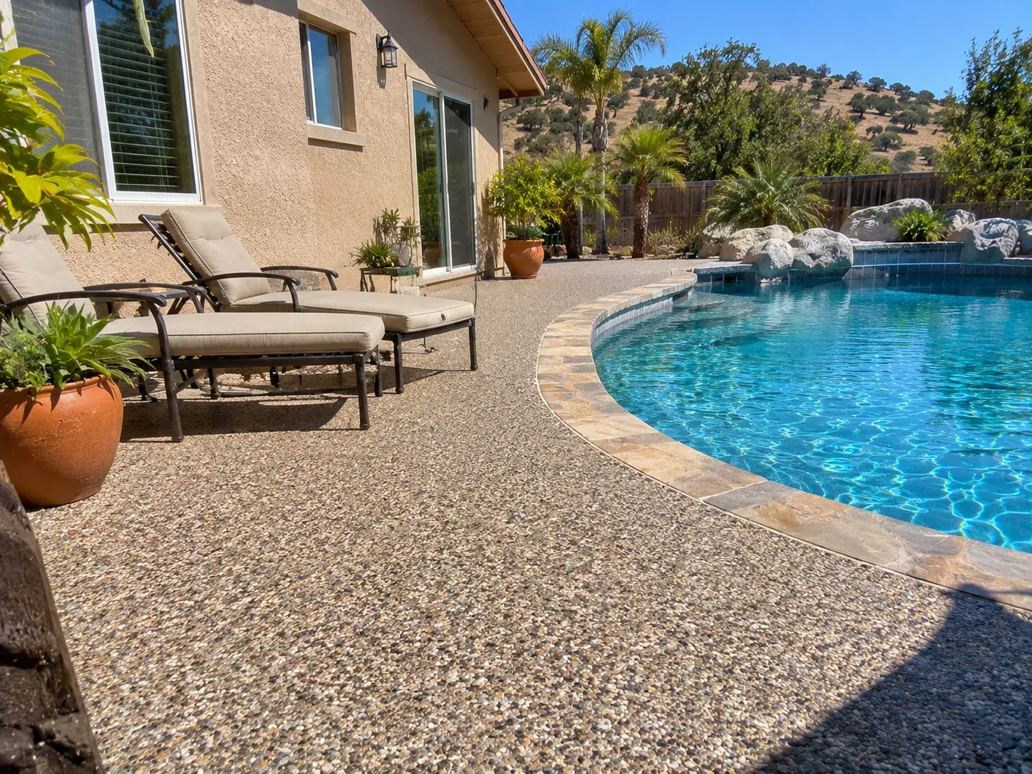 An exposed-aggregate concrete pool deck curving around a blue pool with stone coping, with loungers beside a stucco home and oak-dotted hills behind.