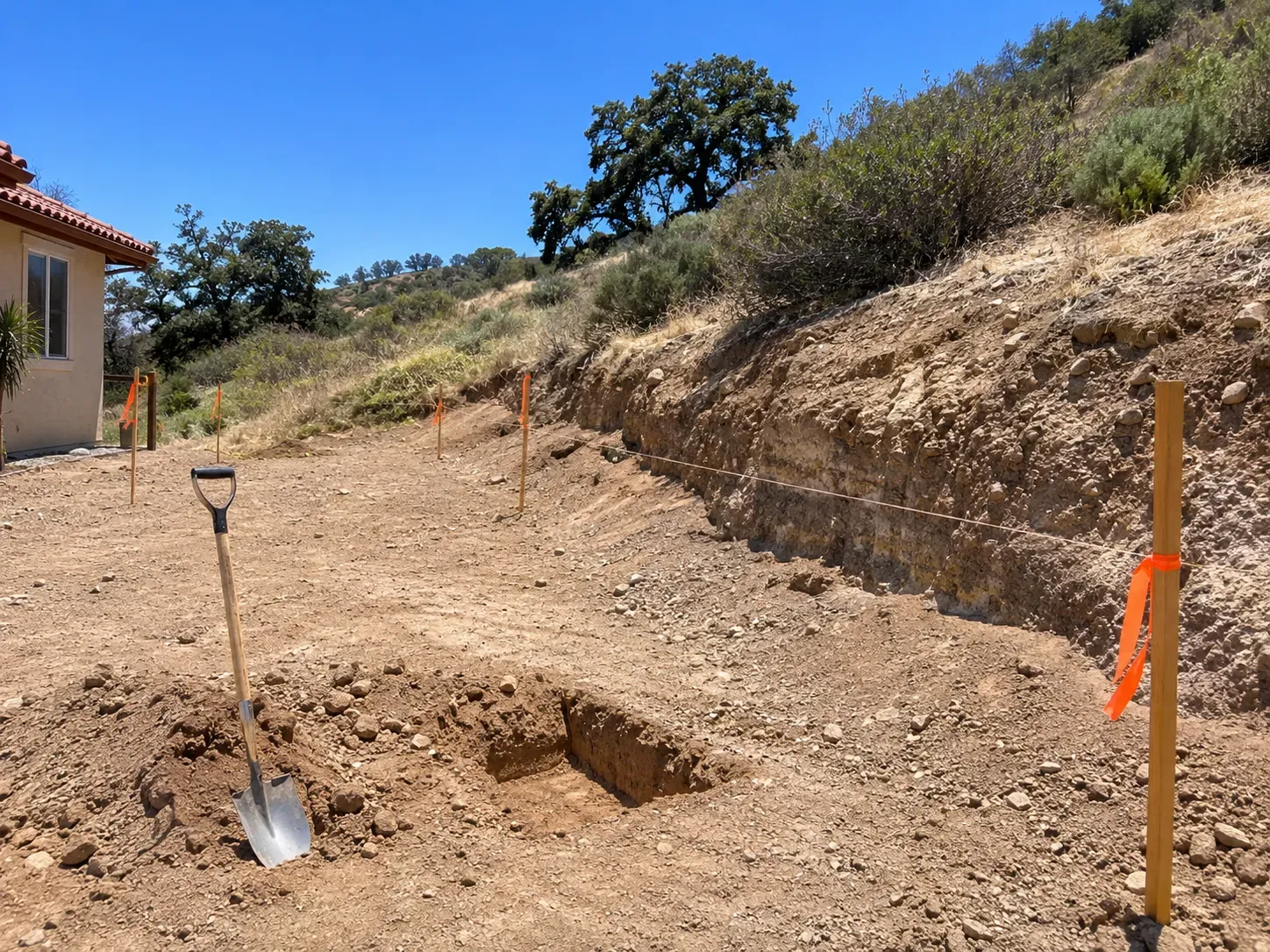 A graded hillside building pad with a cut bank showing the soil layers, survey stakes with orange flagging and a string line, and a test hole dug to check the soil, below chaparral and oaks.