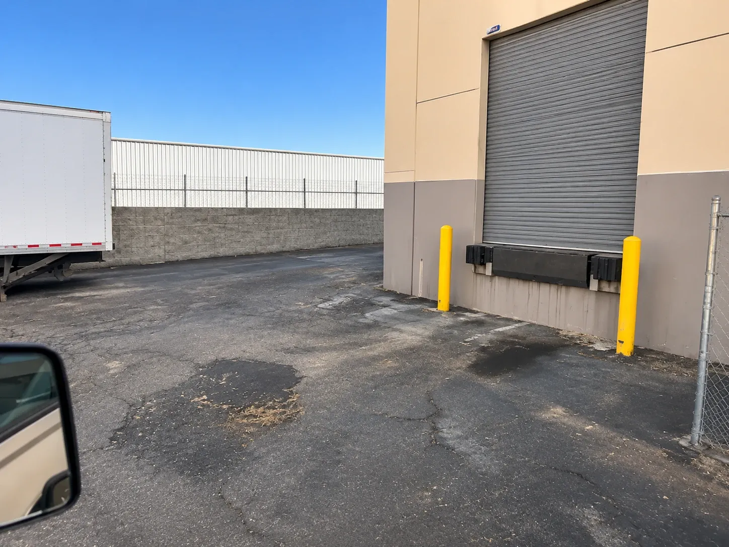 Before: broken, patched asphalt with oil stains in front of a warehouse loading dock door with yellow bollards, a box trailer parked at the left.
