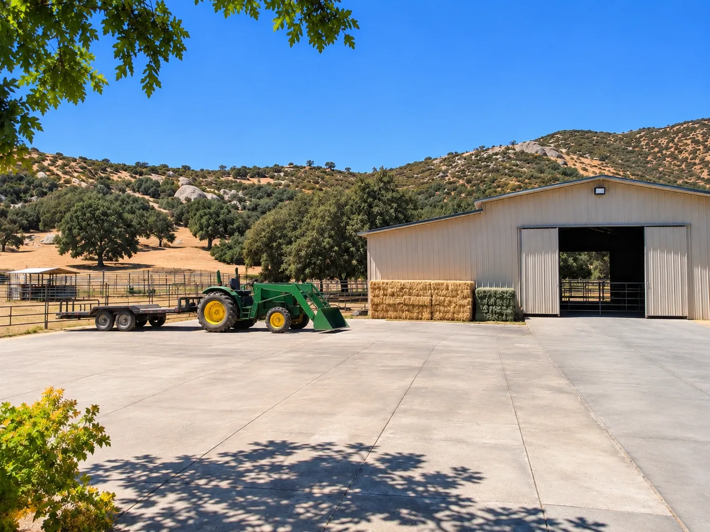 A broad concrete equipment pad and access drive in front of a metal ranch barn, with a tractor and trailer parked on it, hay bales and rail-fenced paddocks below boulder-strewn hills.