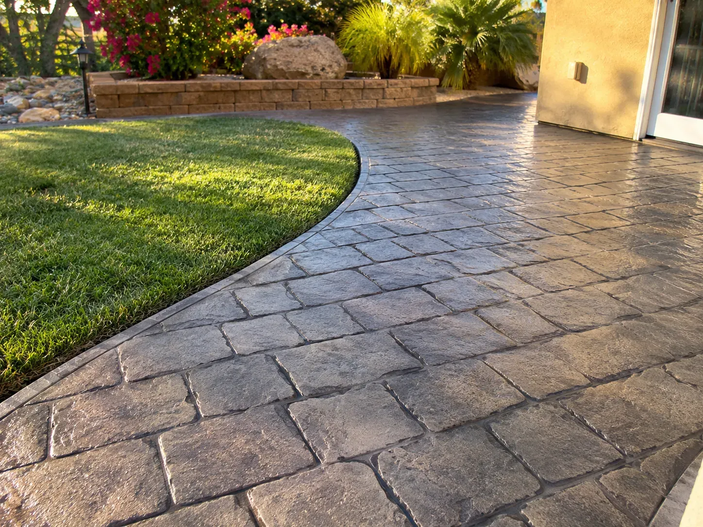 A sealed stamped concrete walkway in a charcoal cobblestone pattern curving cleanly along a lawn edge beside a stucco home, lit by low late-afternoon sun.