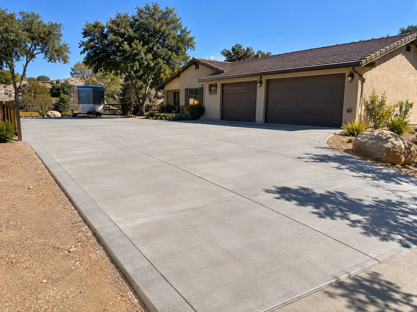 A wide new broom-finished concrete driveway with saw-cut joints and a smooth border band, leading to a ranch home's three-car garage, with a trailer parked on the side area.